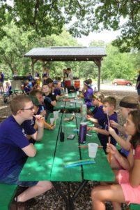 Boy Scouts and Girl Scouts gathered at picnic tables under a shelter during an outdoor activity